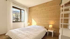 A bedroom with a wooden wall, a bed with white bedding, two bedside lamps, and a window with curtains. - Cozy apartment in Praz-sur-Arly with parking and balcony, near slopes, for 6 people (Praz-sur-Arly)