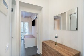 A hallway with a wooden console table, a mirror, and a door leading to a room with a bed and a window.