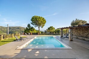 A rectangular swimming pool with a stone wall and a pergola on one side, surrounded by lounge chairs.