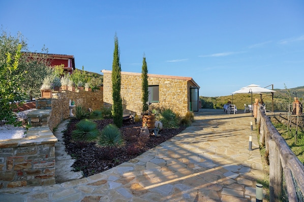 A stone pathway leading to a building with a red roof, surrounded by greenery and a fenced area.