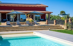 A pool with clear blue water, a red building with a tiled roof, and outdoor seating with umbrellas.