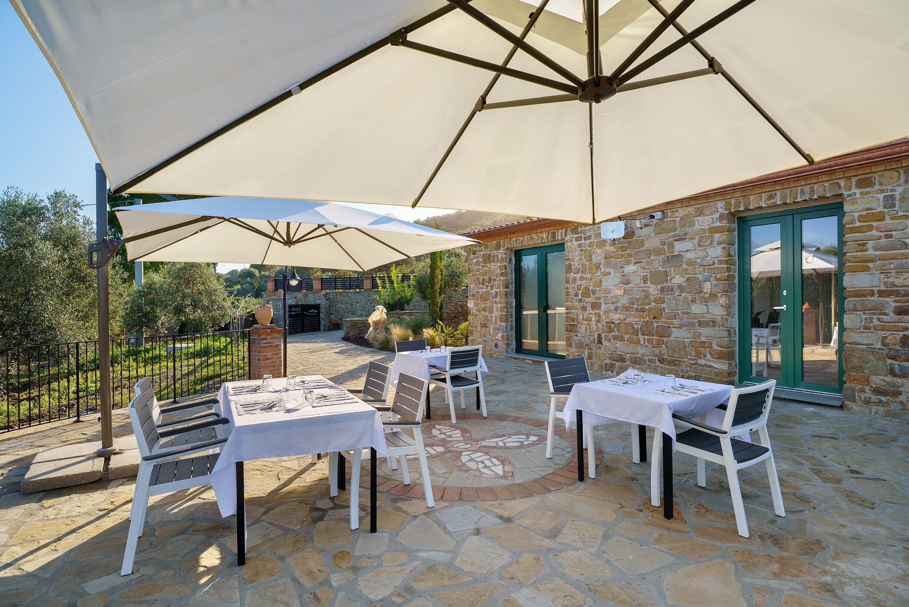 Patio esterno con pavimento in pietra, tavoli e sedie bianche sotto grandi ombrelloni.