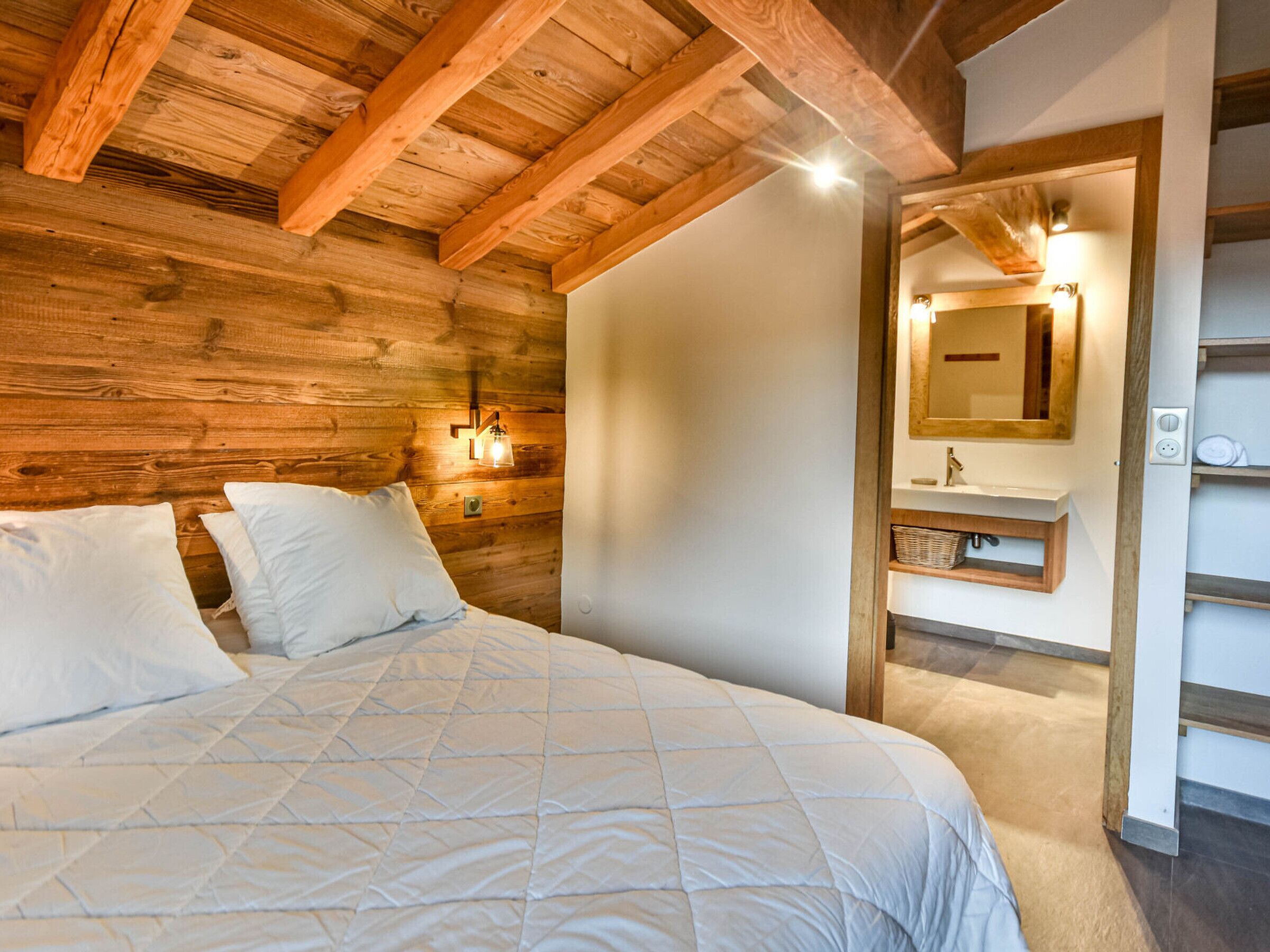Une chambre avec un plafond en bois, un lit avec une literie blanche, un luminaire fixé au mur et un miroir au-dessus d’un lavabo.
