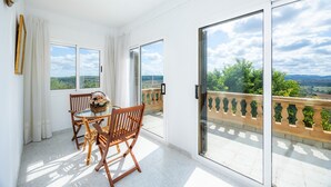 Ein Balkon mit Tisch und Stühlen, Blick auf Grünflächen und Hügel, und ein heller Innenraum.