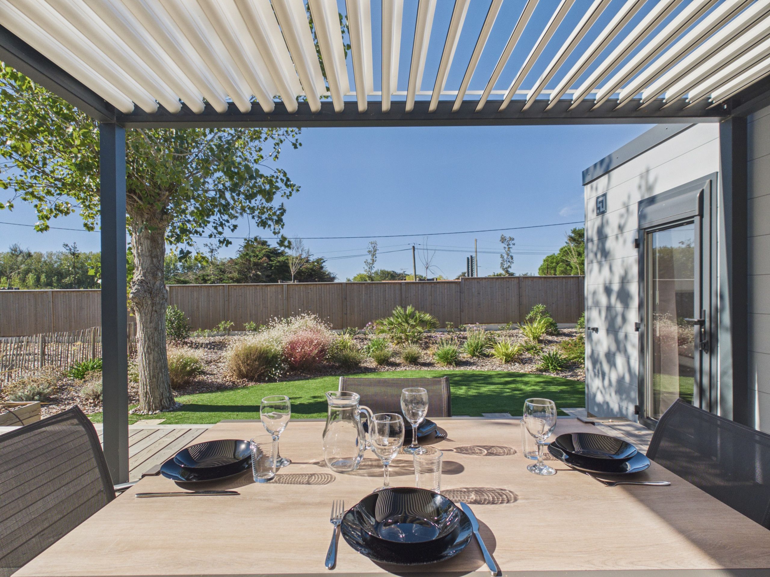 Un espace extérieur aménagé pour les repas, avec une table dressée pour quatre personnes, entouré d’une clôture en bois et d’un jardin.