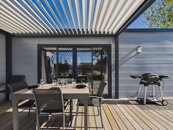 An outdoor patio with a table set for dining, chairs, a grill, and a pergola overhead.