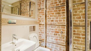 A bathroom with a sink, toilet, and shower area, featuring exposed brick walls and a mirror above the sink. - Experience boutique comfort above The Powell pub in Birchington. Stylish en-suite rooms, luxury touches, and a vibrant pub below—perfect for business, couples, or families. Book your coastal escape today! (Birchington)