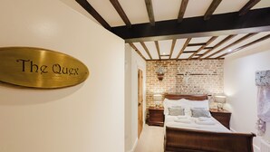 A hotel room with a bed, a wooden headboard, a nightstand with a lamp, a brick wall, and a brass plaque on the wall.