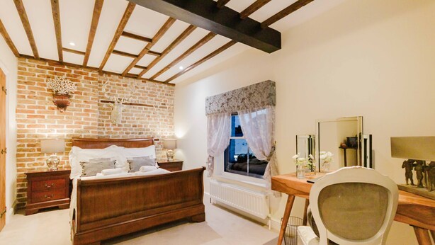 A bedroom with a brick wall, a wooden bed, a desk with a chair, a window with curtains, and a mirror.