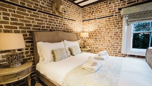 Quarto com parede de tijolos, cama de madeira com roupa de cama branca, mesa de cabeceira com abajur e janela com vista para vegetação.