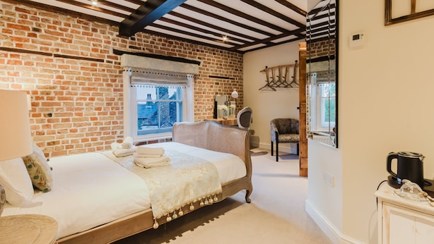 Quarto com parede de tijolos, cama de madeira, janela com vista, uma cadeira e uma mesinha com chaleira.