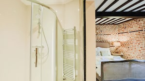 Uma casa de banho moderna com cabine de duche em vidro e sanita separada.  Junto à casa de banho encontra-se um quarto com parede de tijolo, cama e mesa de cabeceira com candeeiro.