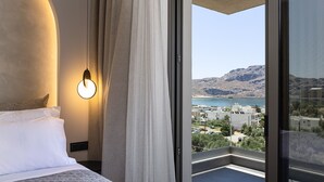 A hotel room with a bed, a nightstand, a wall-mounted light, and a view of a coastal landscape through a large window.