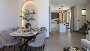 A modern dining area with a round table, grey chairs, and a vase with greenery. A kitchen and living area are visible in the background.