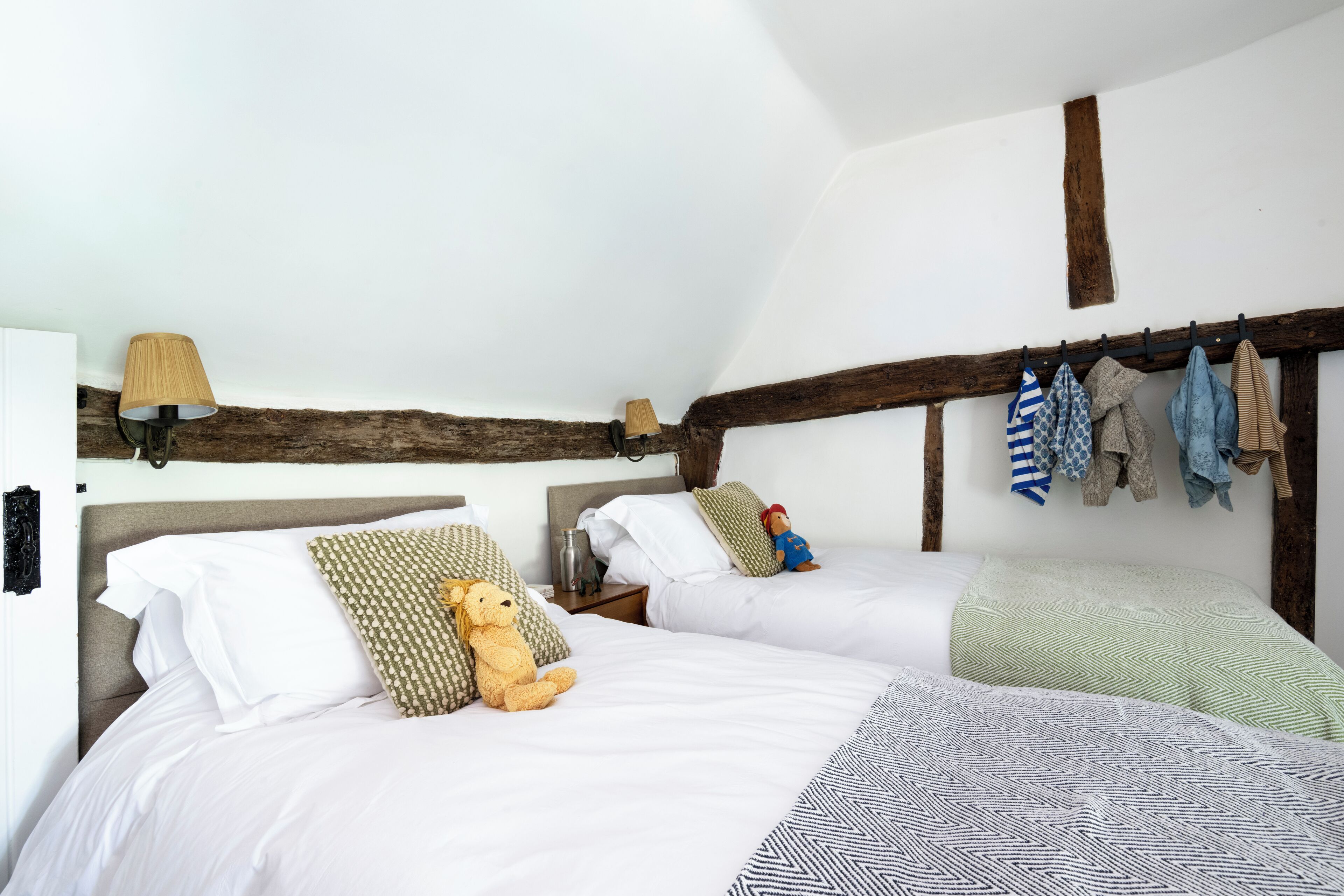 Ein Schlafzimmer mit zwei Betten, Holzbalkendecke, einer Lampe und einem Kleiderständer mit Kleidung.