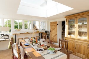 Un espace repas avec une longue table en bois dressée pour le repas, un buffet en bois aux portes vitrées et une lucarne au-dessus.