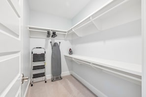 Ein schmaler begehbarer Kleiderschrank mit weißen Wänden, einem Metallbügelbrett, einer Stehleiter und einem kleinen Regal mit einem einzelnen Gegenstand.