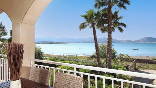 A balcony with a view of a calm sea and mountains, a palm tree, and a wicker chair.