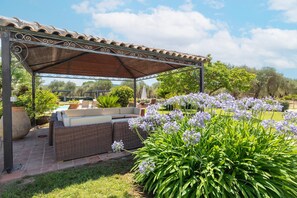 Un espacio de estar techado al aire libre con mobiliario de mimbre y un jardín con flores púrpuras.