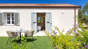 Un bâtiment avec un toit recouvert de tuiles, une porte dont les volets sont ouverts, une petite table avec des chaises et une pelouse bien entretenue agrémentée de fleurs.
