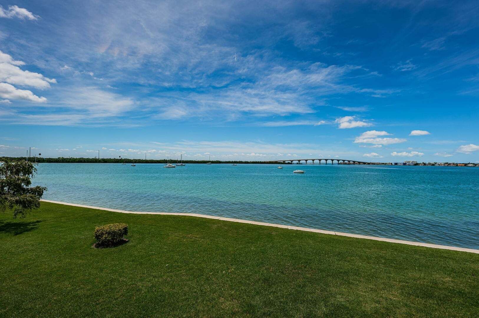 A waterfront view with a bridge, calm water, and a grassy area.