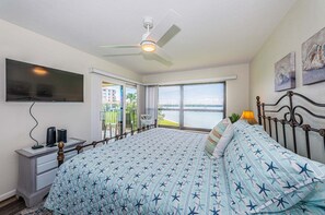 Een slaapkamer met een bed, een plafondventilator, een aan de muur bevestigde televisie en een raam met uitzicht op het water.