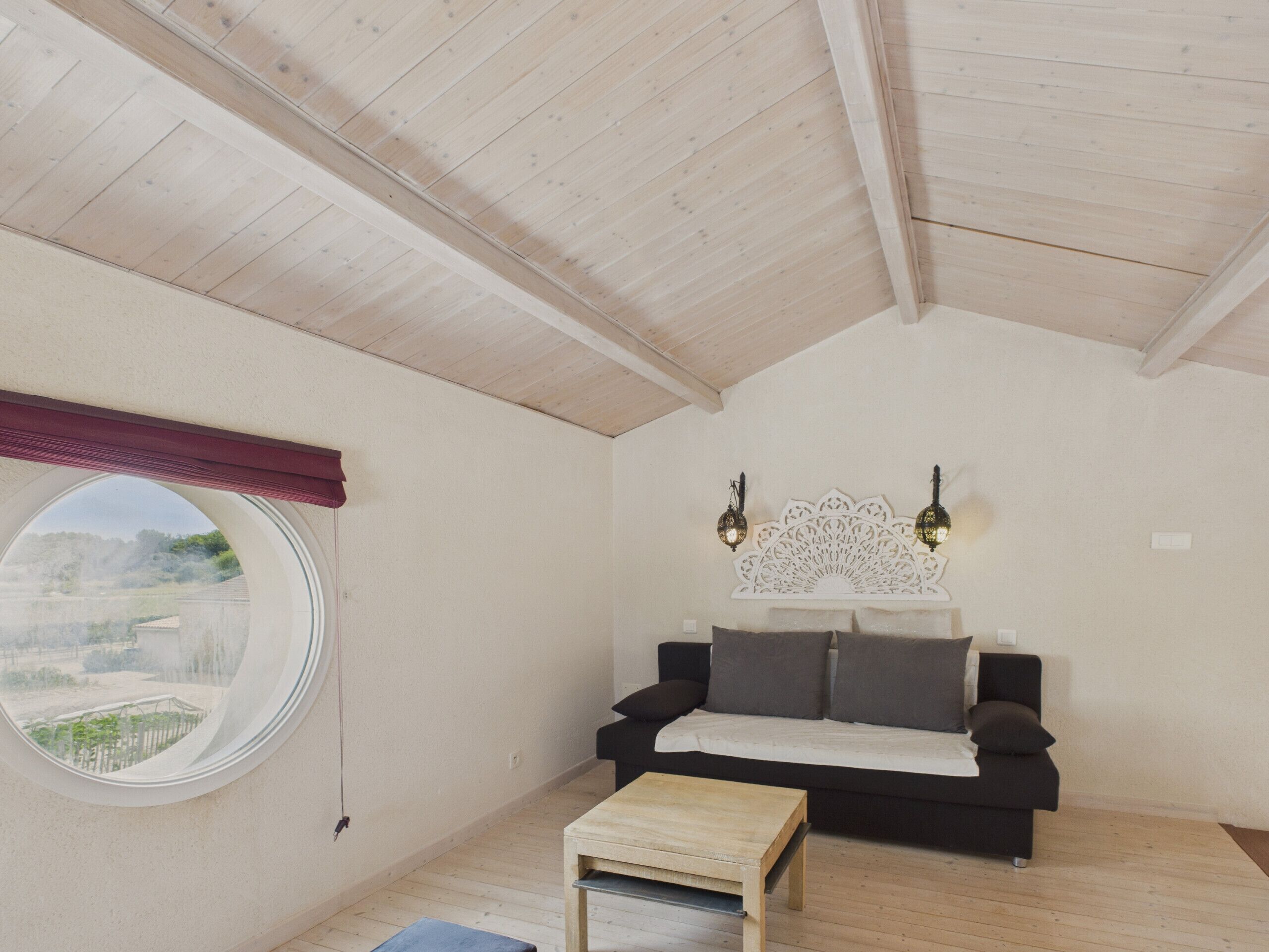 A cozy room with a wooden ceiling, a round window, a black sofa, and two wall-mounted lights.