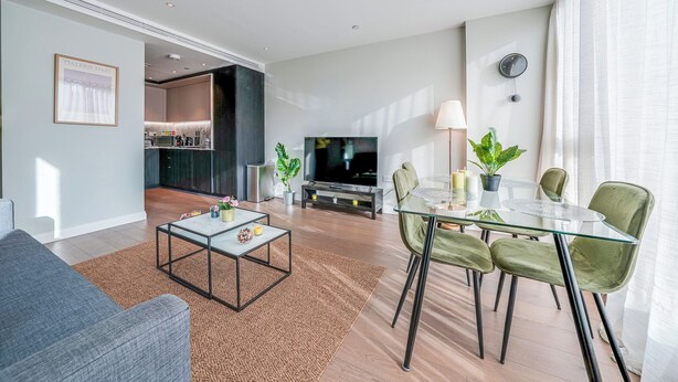 Un salon moderne avec un canapé, une table basse et un coin repas comprenant des chaises et une table en verre. Un téléviseur est fixé au mur, et on aperçoit une cuisine au fond.