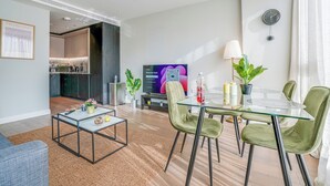 Un salon moderne avec une table à manger en verre, des chaises recouvertes d’un tissu vert, un canapé, une table basse, un téléviseur et, au fond, un coin cuisine.