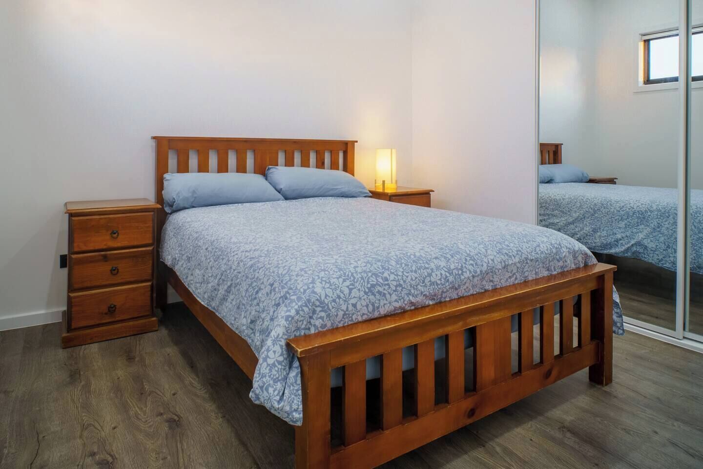 A single bed with a wooden frame and a floral-patterned bedspread, flanked by two bedside tables with lamps, in a room with wooden flooring and a large mirror on the wall.