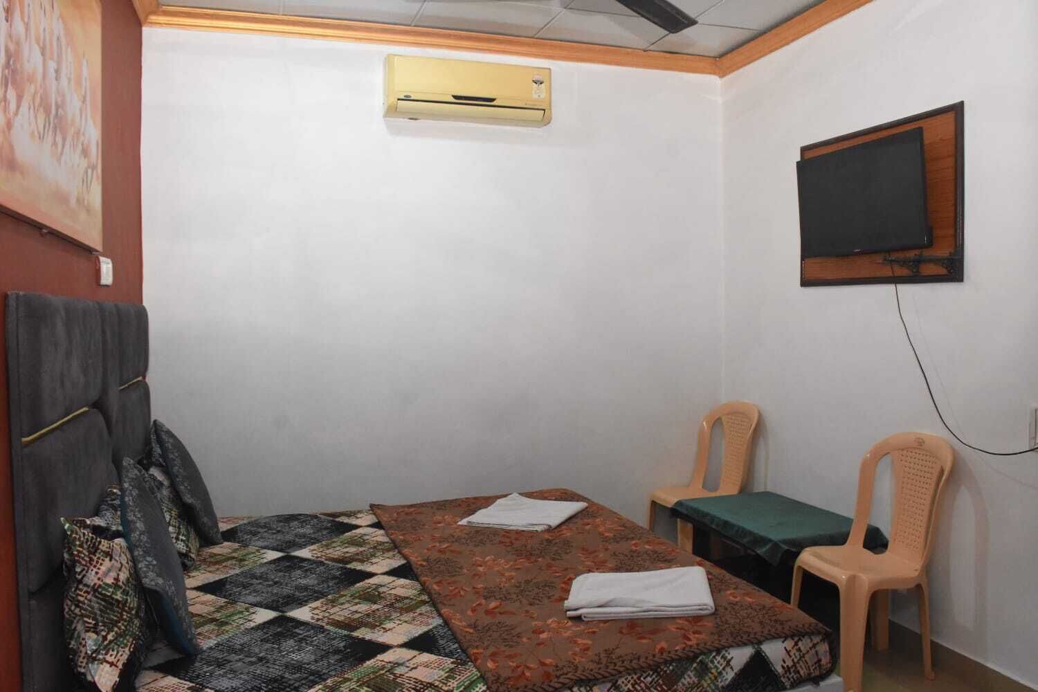 Ein schlichter, spärlich möblierter Raum mit einem Bett, zwei Stühlen, einem an der Wand befestigten Fernseher und einer Klimaanlage.