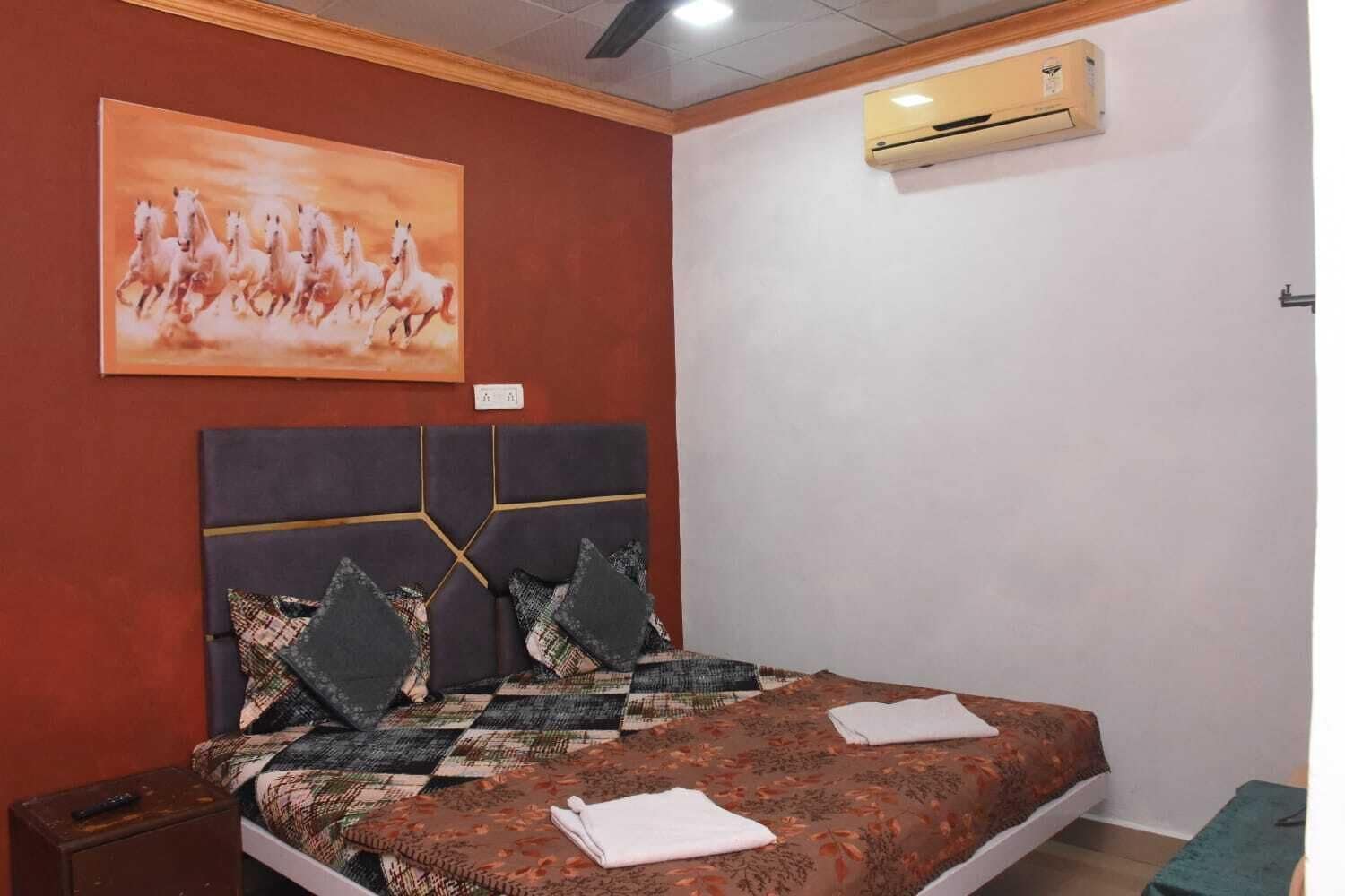 A bedroom with a bed, a painting, an air conditioner, and a nightstand.