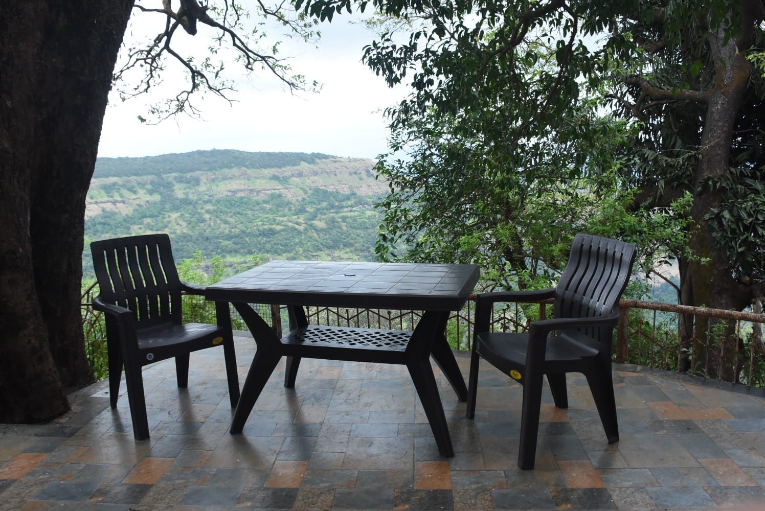 An outdoor seating area with a table and two chairs, surrounded by trees and a view of a hilly landscape.