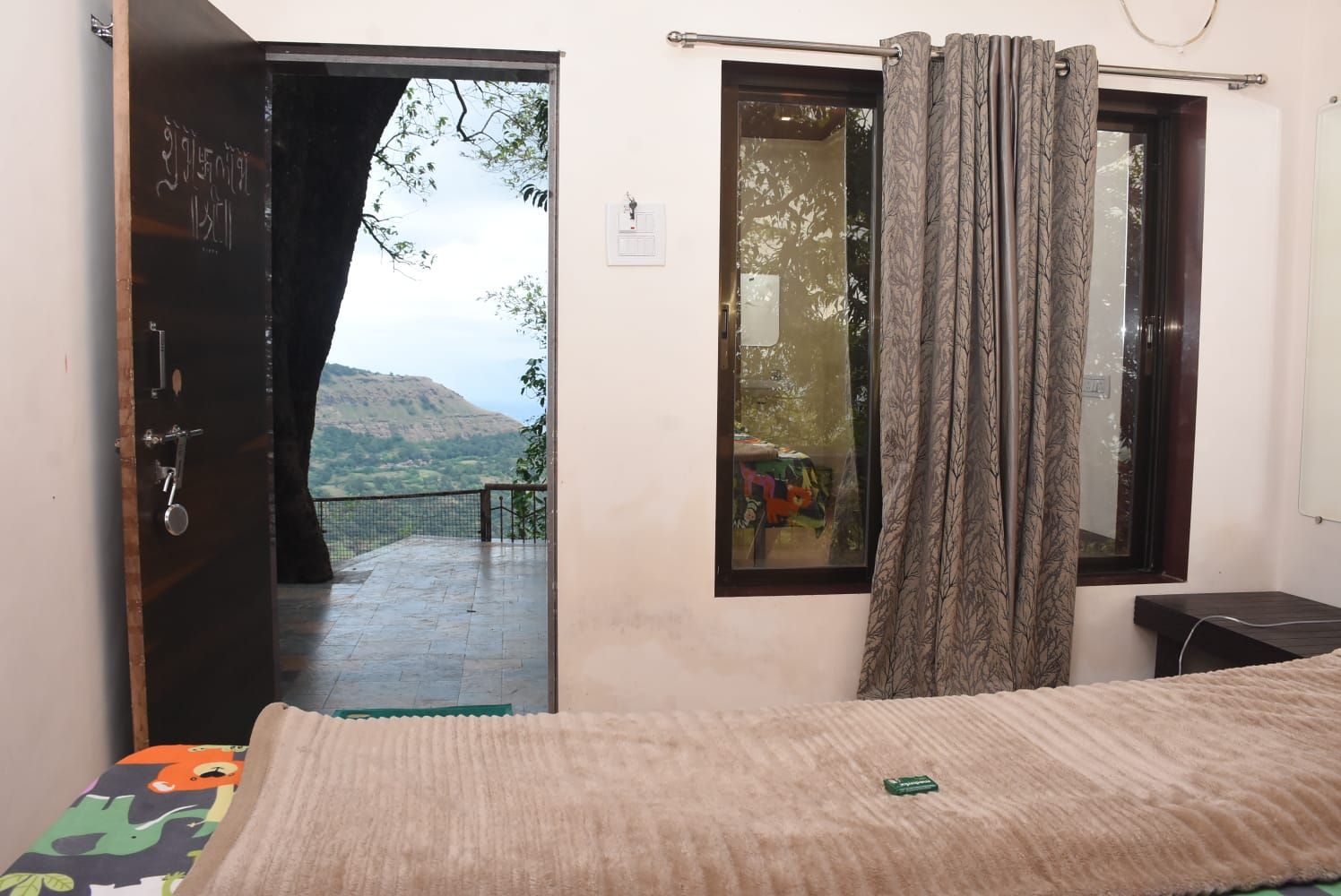 A room with a bed, a window with curtains, a door leading to an outdoor balcony, and a small bedside table.