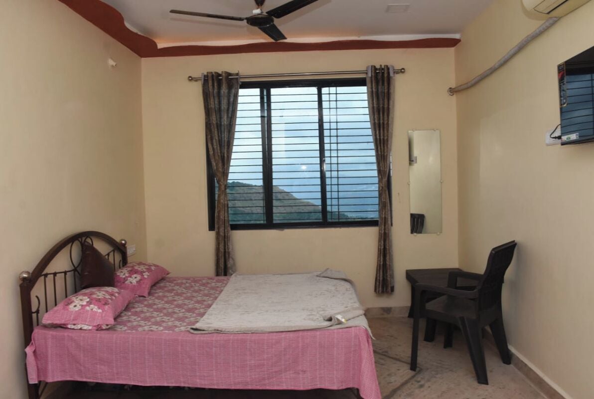 A bedroom with a bed, a desk, a chair, a ceiling fan, a window with curtains, and a wall-mounted air conditioning unit.
