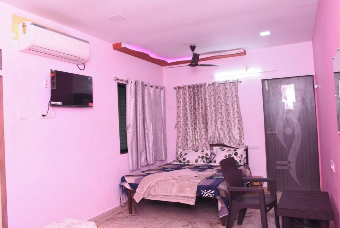 A bedroom with a bed, a desk, a chair, an air conditioner, a ceiling fan, and a television.