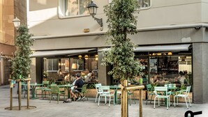 An outdoor cafe with green chairs and tables, surrounded by trees and a building with a large window. - Hotiday Barcelona Colom (Barcelona)