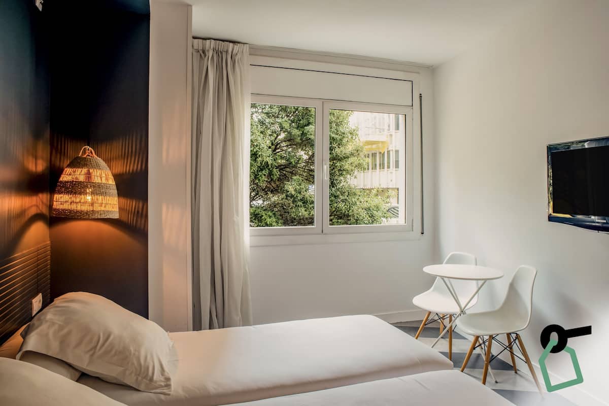 A hotel room with a bed, a small table with chairs, a TV, and a window with curtains.