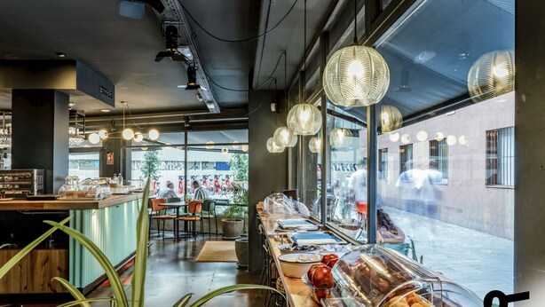 A modern cafe with a long counter, clear display cases, and hanging paper lanterns. - Hotiday Barcelona Colom (Barcelona)
