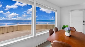 A room with a large window overlooking a beach, a wooden table with chairs, and a potted plant. - 2 BR Unit w/ Mesmerizing Water View @Oak Harbor (Oak Harbor)