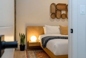 Quarto bem organizado com cama, mesa de cabeceira, luminária, planta em vaso e arte decorativa na parede.
