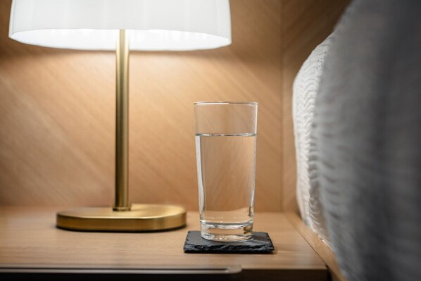 A clear glass of water on a black coaster next to a lamp on a wooden bedside table.