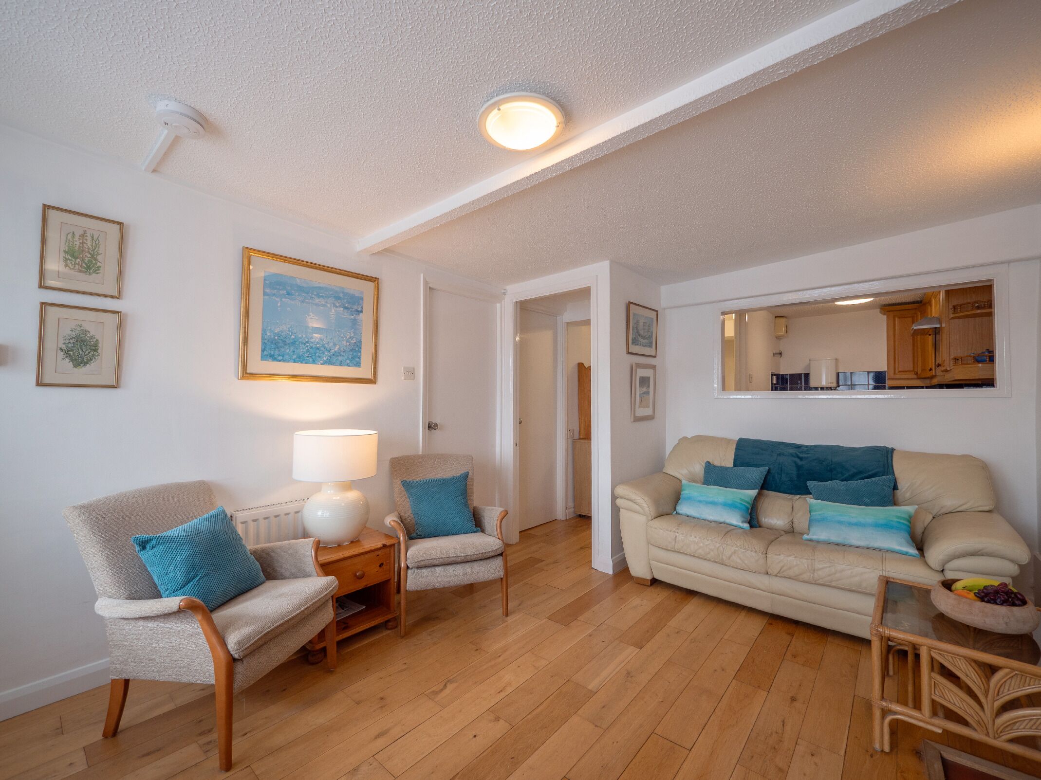 En stue med en beige sofa, to lænestole, et sofabord og en lampe. Væggene er dekoreret med indrammede billeder, der er en loftlampe, og en dør, som fører til et andet rum.