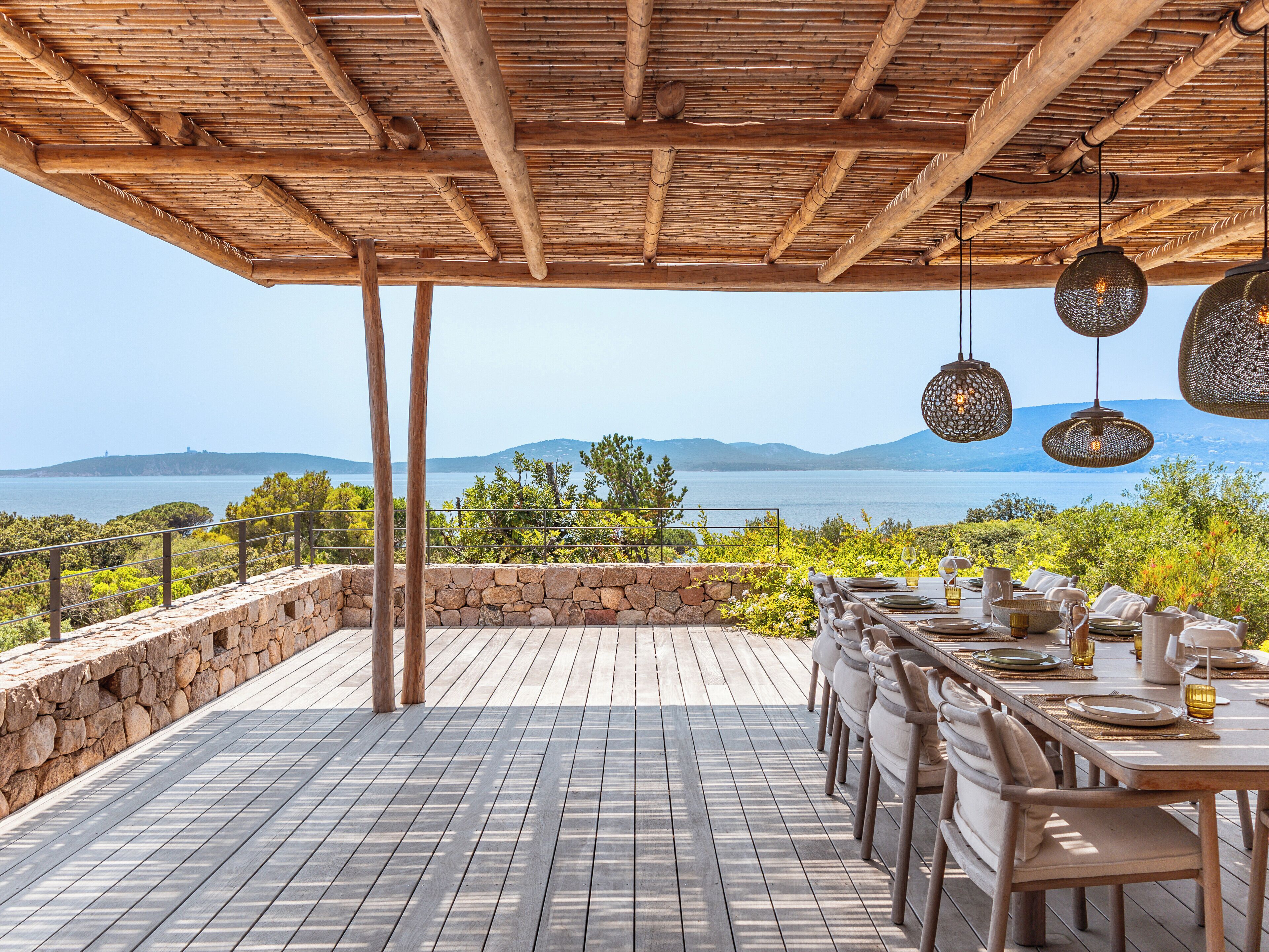 An outdoor dining area with a view of the sea, wooden pergola, and hanging light fixtures.