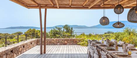 An outdoor dining area with a view of the sea, wooden pergola, and hanging light fixtures.