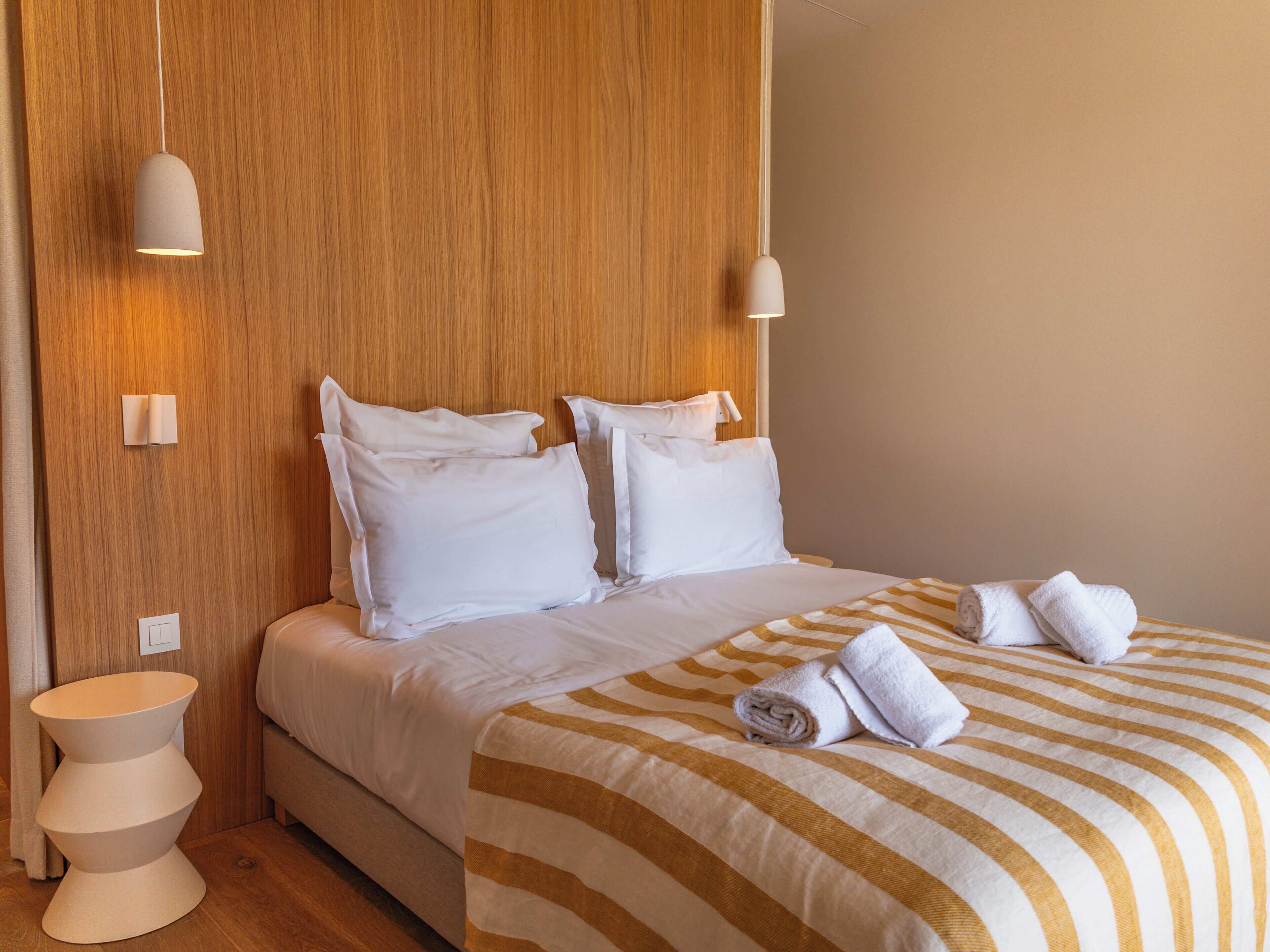 Una cama bien tendida con ropa de cama blanca y mantas a rayas, una mesita de noche con lámpara y un cabecero de madera.