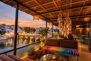 An outdoor seating area with a view of a river and cityscape, featuring a wooden ceiling, hanging dreamcatchers, and potted plants. - Venice View Hotel & Restaurant (Udaipur)