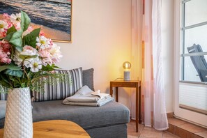 Ett mysigt rum med en grå soffa, ett litet träbord med en lampa, en vas med blommor och ett fönster med gardiner.