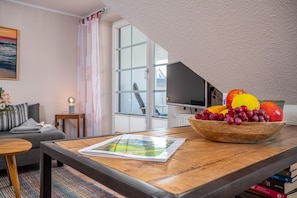 A living room with a wooden table, a bowl of fruit, a sofa, a TV, and a window with curtains. - Störtebekerstraße 9 - Apartment 6 - Comfortable vacation apartment with covered terrace (Zingst)