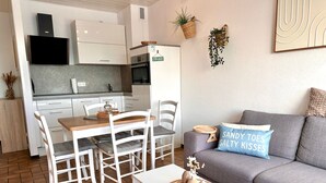 A modern kitchen with white cabinets, a built-in oven, and a refrigerator. There is a dining area with a wooden table and white chairs. A grey sofa with decorative pillows is in the living area.